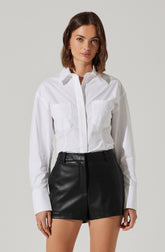 A woman is wearing a white long-sleeve button-up shirt with a collar and two chest pockets. She is also wearing high-waisted black leather shorts with belt loops, a front zipper, and a slightly loose fit.
